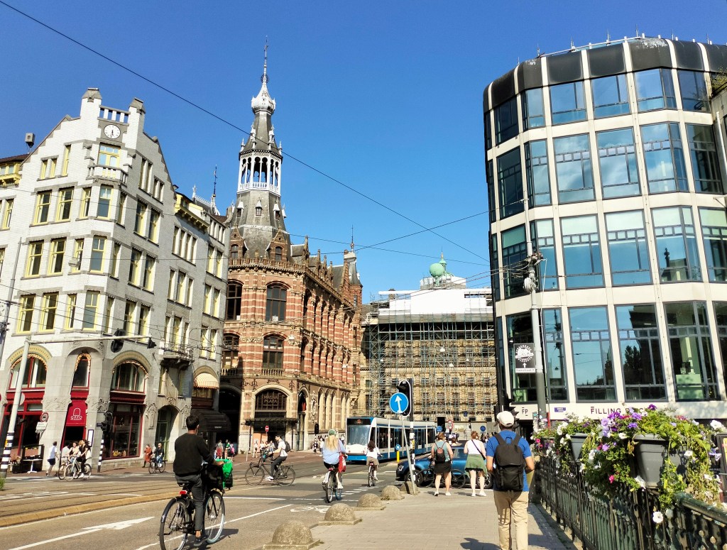 A busy street in Amsterdam with cars, pedestrians, cyclists, and a tram moving by.